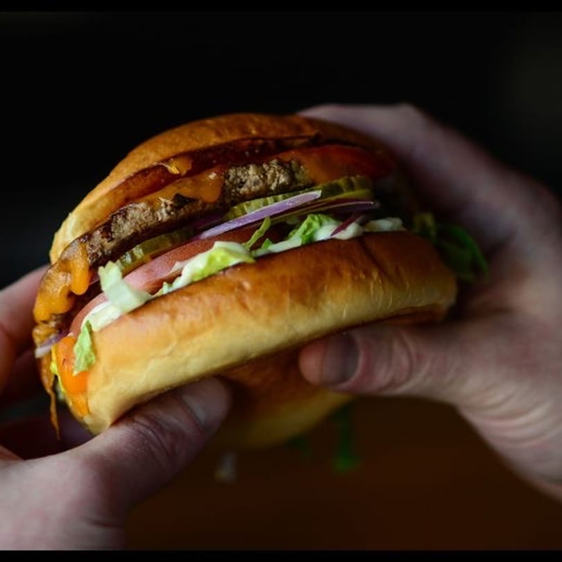 Hands holding a Drip burger with a beef patty, cheese, lettuce, tomato, onions, and pickles.