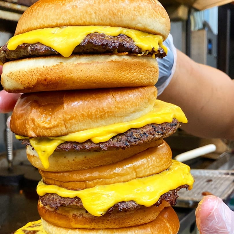 A towering Drip burger with multiple patties and melted cheese, held by a gloved hand.